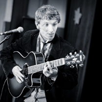 zdjecie wykonawcy - mężczyzna stojący na scenie przy mikrofonie, grający na gitarze