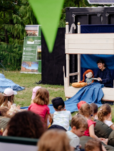 Mini scena na trawie, gdzie Pani persofinikuje postać lalki. Przed nią siedzą dzieci na trawie.