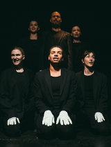 Trzy osoby siedzące obok siebie, w czarnych ubraniech i z białymi rękawiczkami na rękach. Za nimi ich odbicie. Czarne tło.