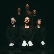 Trzy osoby siedzące obok siebie, w czarnych ubraniech i z białymi rękawiczkami na rękach. Za nimi ich odbicie. Czarne tło.