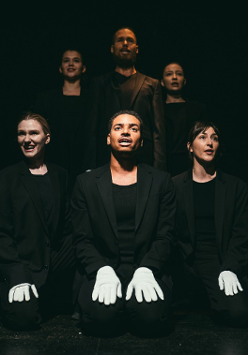 Trzy osoby siedzące obok siebie, w czarnych ubraniech i z białymi rękawiczkami na rękach. Za nimi ich odbicie. Czarne tło.