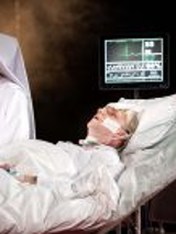 na łóżku szpitalnym leży męzczyzna podłączony do aparatury medycznej, obok stoi siostra zakonna