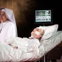 na łóżku szpitalnym leży męzczyzna podłączony do aparatury medycznej, obok stoi siostra zakonna