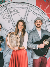 na zdjęciu dwóch mężczyzn i kobieta grający na instrumentach muzycznych na scenie, w tle za nimi duży rysunek pizzy