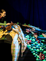 Na zdjęciu dwie kobiety, jedna trzyma pluszową wiewiórkę, druga - dzięcioła; obie aktorki stoją po dwóch stronach półkolistej konstrukcji złożonej z pasów materiału i kolorowych kulek.