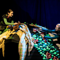 Na zdjęciu dwie kobiety, jedna trzyma pluszową wiewiórkę, druga - dzięcioła; obie aktorki stoją po dwóch stronach półkolistej konstrukcji złożonej z pasów materiału i kolorowych kulek.