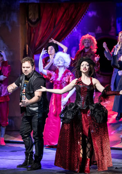 Zdjęcie ze spektaklu - na scenie aktorzy w różnorodnych strojach; na pierwszym planie aktorka w długiej balowej sukni, obok niej mężczyzna w skórzanych spodniach, skórzanej kamizelce i T-shircie.