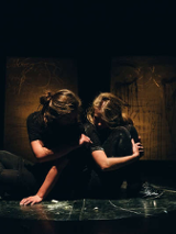 Dwie aktorki siedzące obok siebie na scenie, jedna kobieta dotyka drugą ręką, twarze niewidoczne.