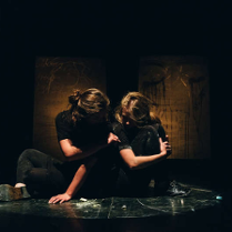 Dwie aktorki siedzące obok siebie na scenie, jedna kobieta dotyka drugą ręką, twarze niewidoczne.