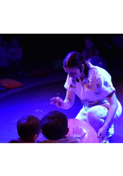 na podłodze obraz nieba i układu słonecznego, wokół siedzą dzieci, przed nimi aktorka ubrana w biały kombinezon, przyklęka na jedno kolano