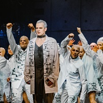 Zdjęcie z przedstawienia: grupa aktorów w białych strojach w różnych pozach na scenie. Na pierwszym planie wyprostowany mężczyzna.