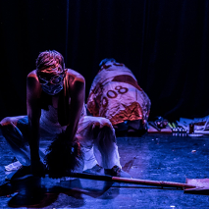 na scenie widoczne dwie postaci, jedna kuca odwrócona tyłem, nakryta jest kolorowym materiałem, druga - mężczyzna, pochyla się podnosząc leżącą na podłodze łopatę