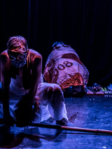 na scenie widoczne dwie postaci, jedna kuca odwrócona tyłem, nakryta jest kolorowym materiałem, druga - mężczyzna, pochyla się podnosząc leżącą na podłodze łopatę