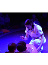 na podłodze obraz nieba i układu słonecznego, wokół siedzą dzieci, przed nimi aktorka ubrana w biały kombinezon, przyklęka na jedno kolano