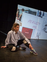 Na zdjęciu młoda kobieta siedzi na podłodze, w tle ściana z różnymi napisami i rysunkami.