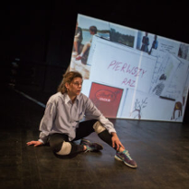 Na zdjęciu młoda kobieta siedzi na podłodze, w tle ściana z różnymi napisami i rysunkami.