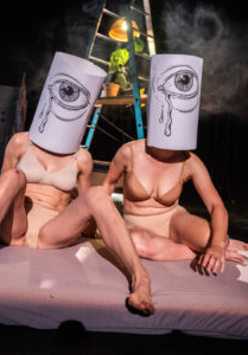 Zdjęcie dwóch siedzących kobiet w strojach bikini, z nałóżonymi na głowy tubami. Na każdej tubie narysowane jest oko, z którego lecą łzy.