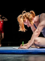 Zdjęcie siedzącej na materacu dziewczyny w sportowym stroju, piszącej długopisem na kartce. W tle stojący mężczyzna w czerwonych stroju sportowym.