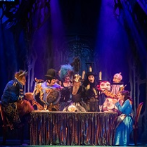 Zdjęcie ze spektaklu w granatowo-czarnych barwach; na pierwszym planie oświetleni aktorzy na scenie siedzący lub stojący wokół stołu.