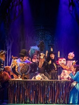 Zdjęcie ze spektaklu w granatowo-czarnych barwach; na pierwszym planie oświetleni aktorzy na scenie siedzący lub stojący wokół stołu.