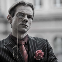 Czarno-białe zdjęcie mężczyzny w koszuli, krawacie i marynarce w paski, z monoklem przy oku. W kieszonce marynarki róża w różowo-szarym kolorze; w tym samym kolorze krawat i koszula mężczyzny.