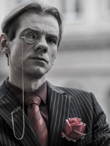 Czarno-białe zdjęcie mężczyzny w koszuli, krawacie i marynarce w paski, z monoklem przy oku. W kieszonce marynarki róża w różowo-szarym kolorze; w tym samym kolorze krawat i koszula mężczyzny.