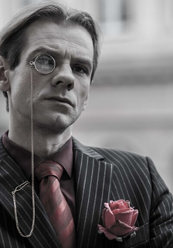 Czarno-białe zdjęcie mężczyzny w koszuli, krawacie i marynarce w paski, z monoklem przy oku. W kieszonce marynarki róża w różowo-szarym kolorze; w tym samym kolorze krawat i koszula mężczyzny.