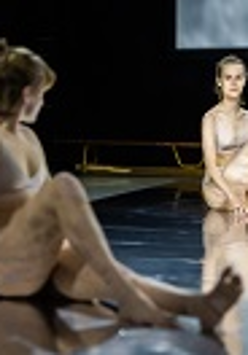 Zdjęcie dwóch młodych kobiet ubranych w biała bieliznę, siedzących na podłodze, każda z podkurczoną nogą. W tle drewniana ławka gimnastyczna.