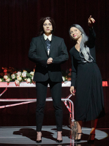 Dwie kobiety, jedna w czarnej sukience, druga w garniturze. Jedna z kobiet wskazuje na coś palcem. Na drugim planie metalowe łóżko na kółkach, na którym leżą białe kwiaty.