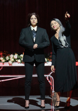 Dwie kobiety, jedna w czarnej sukience, druga w garniturze. Jedna z kobiet wskazuje na coś palcem. Na drugim planie metalowe łóżko na kółkach, na którym leżą białe kwiaty.