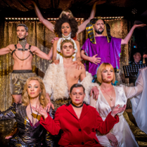 Kilka osób występujących w róznych strojach. Wszyscy patrzą przed siebie, niektórzy mają podniesione ręce. W tle zasłona w złotym kolorze.