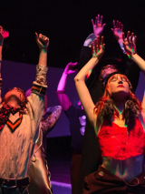 Kilka osób podczas występu. Na pierwszym planie dwie osoby, z podniesionymi rękoma i uniesionymi głowami. Ciemne tło.