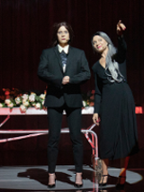 Na pierwszym planie dwie stojące postaci ubrane na czarno, za nimi wysokie metalowe łóżko na kółkach okryte kwiatami