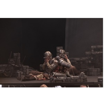 na scenie siedzą tyłem do siebie dwaj mężczyźni ubrani częściowo w zbroje, twarze zakrywają rękami