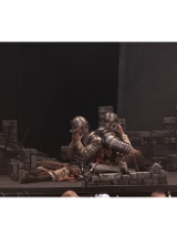 na scenie siedzą tyłem do siebie dwaj mężczyźni ubrani częściowo w zbroje, twarze zakrywają rękami