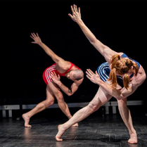 Dwoje aktorów w sportowych strojach na scenie, w takich samych pozach - pochyleni, ze skrzyżowanymi nogami i jedną ręką wyprostowaną ku górze.