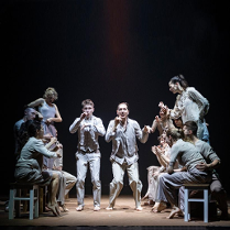 Grupa tancerzy w jasnych ubraniach podczas spketaklu: w środku dwóch mężczyzn stojących na ugiętych nogach, po bokach po kilka osób stojących bokiem lub siedzących na taboretach.