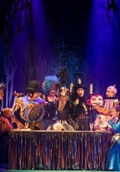 Zdjęcie ze spektaklu w granatowo-czarnych barwach; na pierwszym planie oświetleni aktorzy na scenie siedzący lub stojący wokół stołu.