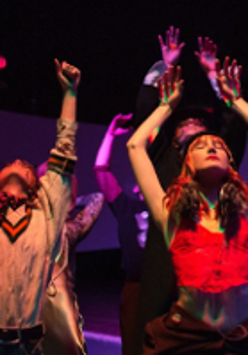 Kilka osób podczas występu. Na pierwszym planie dwie osoby, z podniesionymi rękoma i uniesionymi głowami. Ciemne tło.