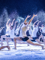 Tancerze na scenie w tanecznych pozach, wszyscy ubrani w białe koszule i czarne, krótkie spodenki. Wokół nich drobny pył przypominający śnieg.