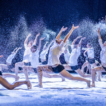 Tancerze na scenie w tanecznych pozach, wszyscy ubrani w białe koszule i czarne, krótkie spodenki. Wokół nich drobny pył przypominający śnieg.