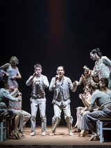 Grupa tancerzy w jasnych ubraniach podczas spketaklu: w środku dwóch mężczyzn stojących na ugiętych nogach, po bokach po kilka osób stojących bokiem lub siedzących na taboretach.