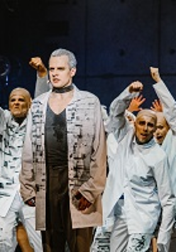 Zdjęcie z przedstawienia: grupa aktorów w białych strojach w różnych pozach na scenie. Na pierwszym planie wyprostowany mężczyzna.