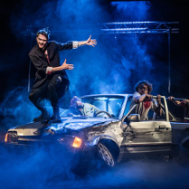 Zdjęcie przedstawiające trzy osoby wysiadające ze zniszczonego samochodu oraz jednego mężczyznę stojącego na masce samochodu.