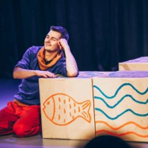 Uśmiechnięty mężczyzna klęcząc podpiera się o drewniane pudło z rusunkiem ryby, obok stoi kolejne pudło z rysunkiem fal.