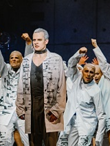 Zdjęcie z przedstawienia: grupa aktorów w białych strojach w różnych pozach na scenie. Na pierwszym planie wyprostowany mężczyzna.