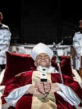 Mężczyzna ubrany w szaty papieskie leży na katafalku, pod głową ma purpurową poduszkę; za nim stoją dwaj mężczyźni w komżach.