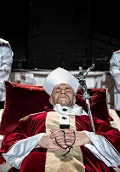 Mężczyzna ubrany w szaty papieskie leży na katafalku, pod głową ma purpurową poduszkę; za nim stoją dwaj mężczyźni w komżach.