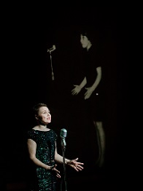 Kobieta w czarnej sukience stoi przed mikrofonem, za nią zdjęcie innej kobiety stojącej przed mikrofonem na czarnym tle.