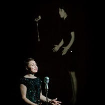 Kobieta w czarnej sukience stoi przed mikrofonem, za nią zdjęcie innej kobiety stojącej przed mikrofonem na czarnym tle.